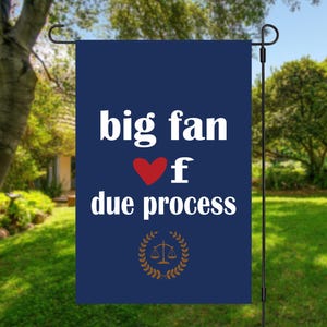 May include: A navy blue garden flag with the text "big fan of due process" in white, a red heart, and a gold scale of justice emblem. The flag is hanging in a garden.