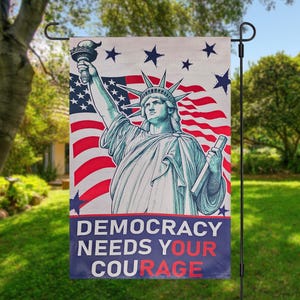 May include: A garden flag featuring the Statue of Liberty with the American flag in the background. The flag has the text "DEMOCRACY NEEDS YOUR COURAGE" in white and red lettering on a blue banner.