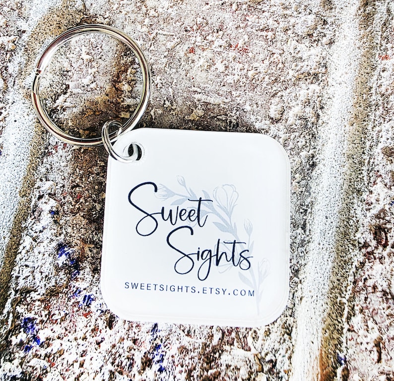 May include: A white square keychain with a silver keyring. The keychain features the words "Sweet Sights" in a script font, with a floral design. The website address SWEETSIGHTS.ETSY.COM is also printed on the keychain.