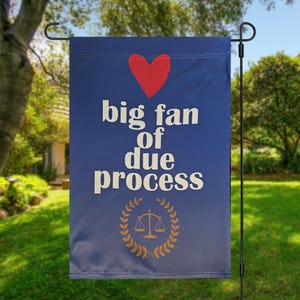 May include: A blue garden flag displaying the words "big fan of due process" in white, a red heart, and a gold scale of justice emblem. The flag is suspended on a black metal stand in a garden.