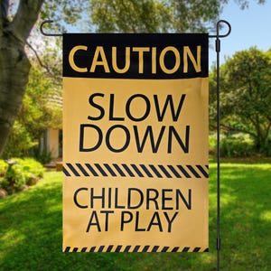 May include: A yellow and black garden flag displaying the text "CAUTION SLOW DOWN CHILDREN AT PLAY". The flag is suspended from a black metal garden stake, set in a green lawn.