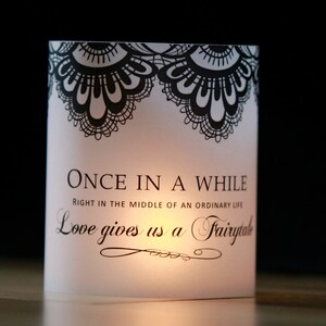 Op de afbeelding: Witte kaarshouder met zwart kantontwerp en de tekst "ONCE IN A WHILE RIGHT IN THE MIDDLE OF AN ORDINARY LIFE Love gives us a Fairytale".