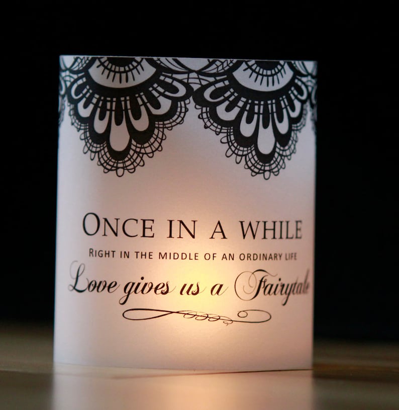 Op de afbeelding: Witte kaarshouder met zwart kantontwerp en de tekst "ONCE IN A WHILE RIGHT IN THE MIDDLE OF AN ORDINARY LIFE Love gives us a Fairytale".