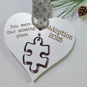 May include: A heart-shaped ornament with the words "You were Our missing piece" and "Adoption 2025" printed on it. A puzzle piece is cut out of the ornament. The ornament is white with a silver lace ribbon.