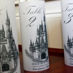 May include: Three white table number cards with black calligraphy. Each card features a black and white illustration of Cinderella's castle and the text "Table" followed by the number 1, 2, or 3. The bottom of each card has the text "Once in a while, right in the middle of an ordinary life, love gives us a fairytale."