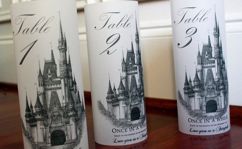 May include: Three white table number cards with black calligraphy. Each card features a black and white illustration of Cinderella's castle and the text "Table" followed by the number 1, 2, or 3. The bottom of each card has the text "Once in a while, right in the middle of an ordinary life, love gives us a fairytale."