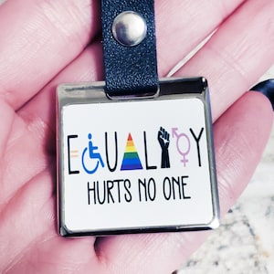 Op de afbeelding: Een zilveren sleutelhanger met een zwart leren bandje. De sleutelhanger heeft een vierkante metalen hanger met een witte achtergrond en de tekst "EQUALITY HURTS NO ONE". De hanger heeft ook een regenboog, een vuist, een rolstoel symbool en een transgender symbool.