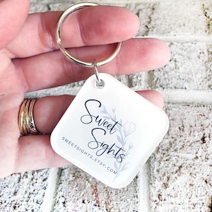 May include: A white, square-shaped keyring with a silver ring. The keyring features the text "Sweet Sights" in a script font, with a floral design. The Etsy shop URL is also printed on the keyring.