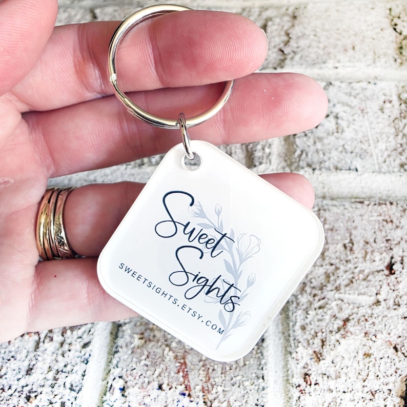 May include: A white, square-shaped keyring with a silver ring. The keyring features the text "Sweet Sights" in a script font, with a floral design. The Etsy shop URL is also printed on the keyring.