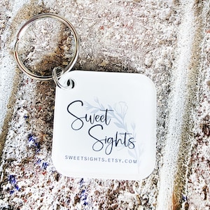 May include: A white square keychain with a silver keyring. The keychain features the words "Sweet Sights" in a script font, with a floral design. The website address SWEETSIGHTS.ETSY.COM is also printed on the keychain.