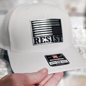 Könnte beinhalten: Weiße Trucker-Mütze mit einem silbernen Aufnäher mit einem amerikanischen Flaggen-Design und dem Wort „RESIST“. Die Mütze hat einen Mesh-Rücken und einen gebogenen Schirm. Die Richardson 112 Mütze ist verstellbar.