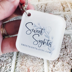 May include: A white, square keyring with rounded corners. The keyring displays the text "Sweet Sights" in a script font, with a floral design and the website address sweetsights.etsy.com.