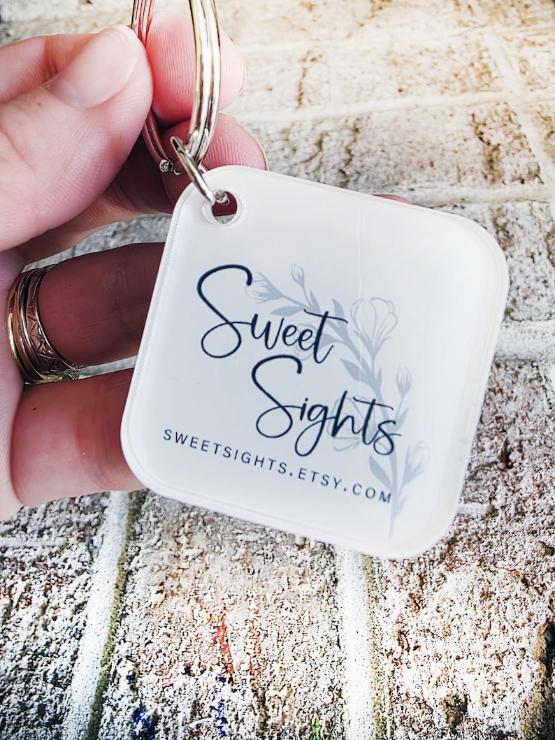 May include: A white, square keyring with rounded corners. The keyring displays the text "Sweet Sights" in a script font, with a floral design and the website address sweetsights.etsy.com.