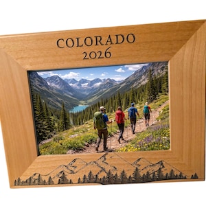 May include: Wooden picture frame with a scenic landscape photo of hikers in the mountains. The frame is light brown with the word "COLORADO" and the year "2026" engraved at the top. A mountain and tree design is etched at the bottom.