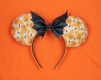 May include: Halloween-themed headband with Mickey Mouse pumpkin ears and a black bow. The ears are patterned with orange pumpkins and bats on a cream background. The headband is orange and black.