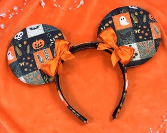 May include: Halloween-themed mouse ear headband with black and orange patchwork design. The ears feature pumpkins, ghosts, and bats. The headband has orange bows and is set against an orange background with decorative pearls.