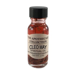 Op de afbeelding: Een klein glazen flesje met een zwarte dop gevuld met een rode vloeistof en kruiden. Het etiket vermeldt "The Apothecary Collection Cleo May Spiritual Oil www.TheApothecaryCollection.com NET WT. .5 oz"