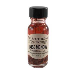 Op de afbeelding: Een klein glazen flesje met een zwarte dop gevuld met een rode vloeistof en kruiden. Het etiket vermeldt "The Apothecary Collection Kiss Me Now Spiritual Oil www.TheApothecaryCollection.com NET WT. .5 oz"