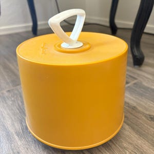 May include: A cylindrical, mustard-yellow container with a white handle. The handle is triangular and attached to the top of the container. The container is sitting on a wooden floor. The background includes a black chair.