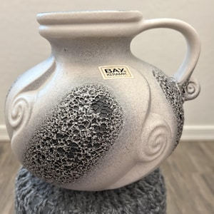 May include: A white and gray ceramic vase with a handle. The vase has a textured, speckled design with dark gray accents and a label that reads "BAY. KERAMIK."