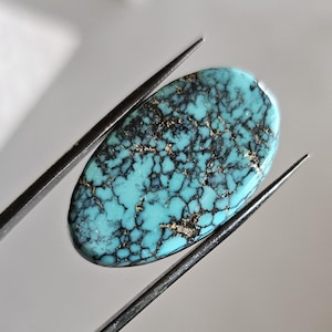 Puede incluir: Gema de turquesa ovalada con una matriz de vetas marrón oscuro y doradas. La piedra pulida está sujeta por unas pinzas metálicas sobre un fondo neutro. La turquesa tiene un color azul-verdoso vibrante.