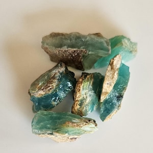 May include: A collection of raw, unpolished mineral stones featuring a translucent teal and blue color palette. Each stone displays a rough, jagged texture with earthy, tan-colored crusts along the edges, showcasing natural variations in shape.