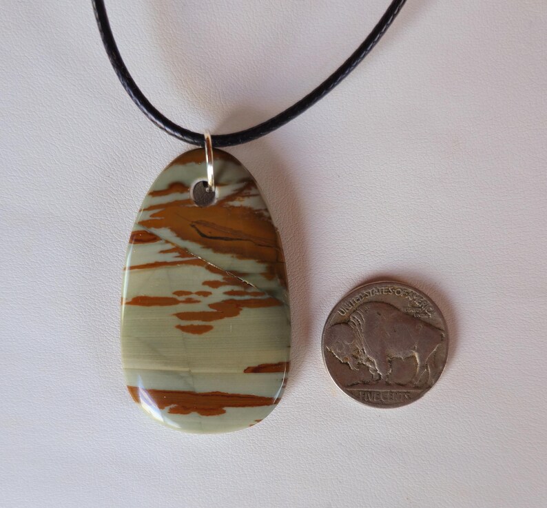 May include: A brown and beige stone pendant with a marbled pattern hangs from a black leather cord. The pendant is oval shaped and has a silver bail.