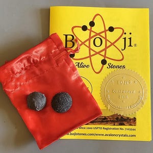 Puede incluir: Dos piedras Boji oscuras y texturizadas descansan en una bolsa roja con cordón, junto a un folleto amarillo. El folleto presenta el texto "Boji Alive Stones" con un símbolo de átomo estilizado y un sello dorado.