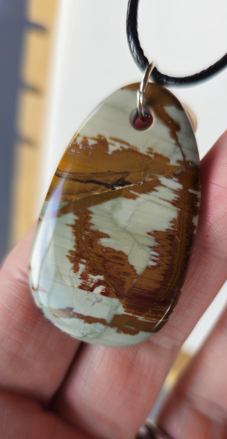 May include: A brown, white, and green oval pendant with a black leather cord. The pendant has a natural stone look with swirls of color.
