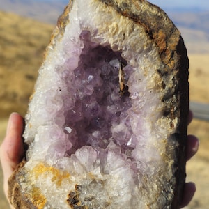 May include: A natural amethyst geode, displaying a cavity filled with sparkling purple crystals. The outer shell is a rough, brown rock, with a white and yellow crystalline border. The geode is held in a hand, with a blurred outdoor background.