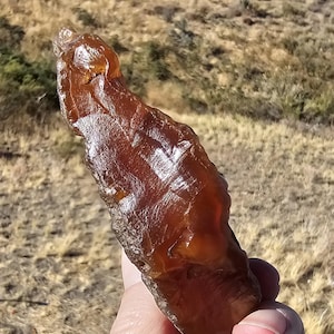 May include: A hand holds a large, amber-colored stone with a glossy surface. The stone has a rough, irregular shape and appears to be translucent, with light passing through it. The background is a blurred view of a dry, grassy landscape.