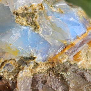 Puede incluir: Primer plano de una piedra preciosa de ópalo en bruto. La piedra presenta secciones translúcidas azules y blancas con áreas marrones y amarillas. La superficie rugosa del ópalo es visible, mostrando su textura natural y variaciones de color.