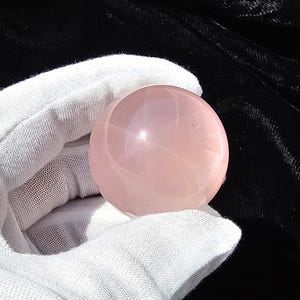May include: A smooth, spherical rose quartz crystal ball. The polished sphere is a soft pink color and held in a white gloved hand against a black background. The crystal ball is a popular item in the metaphysical and spiritual community.
