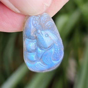 May include: A teardrop-shaped labradorite pendant featuring a detailed relief carving of a fish swimming alongside bamboo stalks. The stone exhibits a vibrant, iridescent blue flash across its polished surface, highlighting the intricate textures of the scales and the segmented bamboo design.