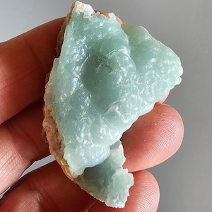 May include: A piece of raw mineral with a light blue, crystalline structure. The mineral has a rough, textured surface and is held in a hand. The edges of the mineral are a mix of white, tan, and pink.