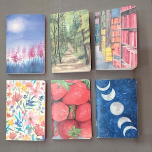 May include: Six different watercolor designs on notebook covers. The designs include a night sky with a moon, a forest path, a bookshelf, a floral pattern, strawberries, and a night sky with crescent moons.