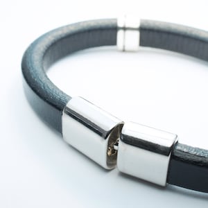 K&ouml;nnte beinhalten: Ein schwarzes Lederarmband mit silbernen Metallverschl&uuml;ssen. Das Armband ist offen und bereit zum Tragen.