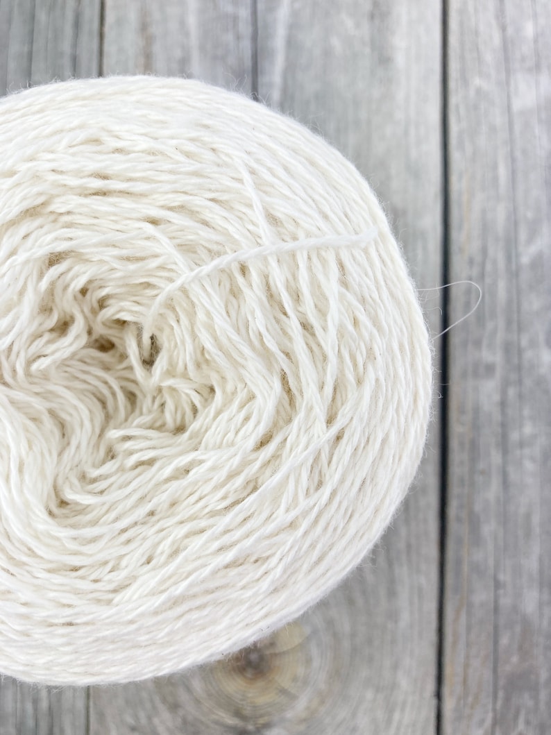 May include: A skein of white yarn, wound into a ball shape, on a wooden surface.