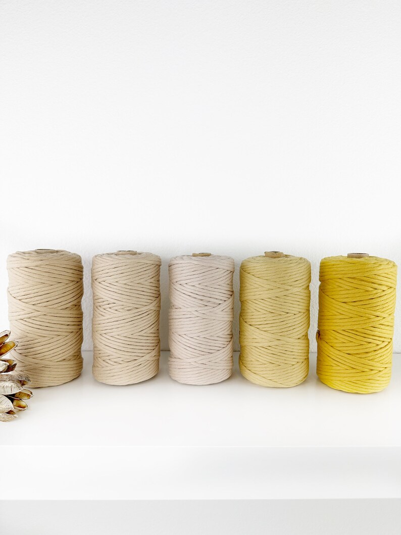 Puede incluir: Cinco carretes de cuerda de algod&oacute;n en varios tonos de beige y amarillo. Los carretes est&aacute;n dispuestos en una fila de izquierda a derecha, con el tono beige m&aacute;s claro a la izquierda y el tono amarillo m&aacute;s oscuro a la derecha.