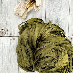 May include: A large bundle of olive green silk ribbon, also known as silk yarn or silk ribbon yarn, is shown on a light wood background. The silk ribbon is made of thin strips of silk fabric that have been frayed and twisted together.
