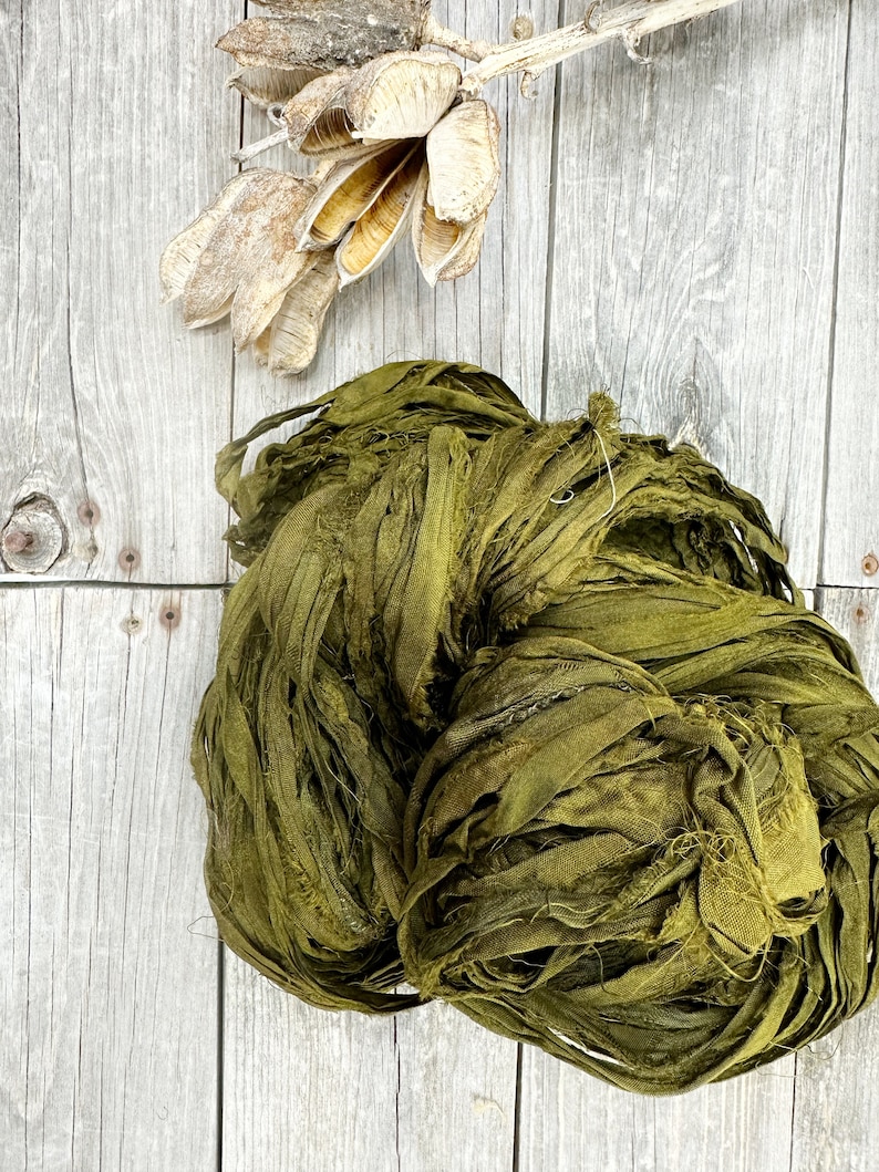 May include: A large bundle of olive green silk ribbon, also known as silk yarn or silk ribbon yarn, is shown on a light wood background. The silk ribbon is made of thin strips of silk fabric that have been frayed and twisted together.