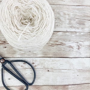 May include: A ball of white yarn sits on a whitewashed wooden surface with a pair of black scissors.
