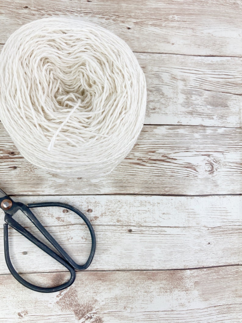 May include: A ball of white yarn sits on a whitewashed wooden surface with a pair of black scissors.