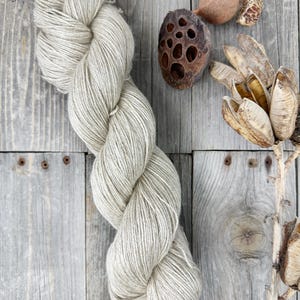 May include: A skein of light gray yarn is the focal point of this image, with a natural, textured appearance. The yarn is twisted and coiled, suggesting it's ready for knitting or crocheting. Dried seed pods and other natural elements are arranged around the yarn, creating a rustic, earthy aesthetic.