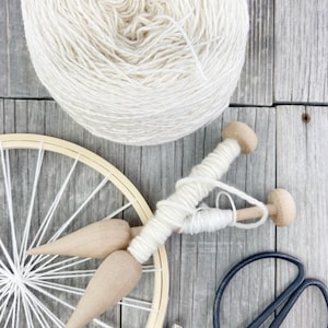May include: A ball of white yarn, a wooden loom with white yarn wrapped around it, two wooden weaving tools, and a pair of black scissors on a wooden surface.