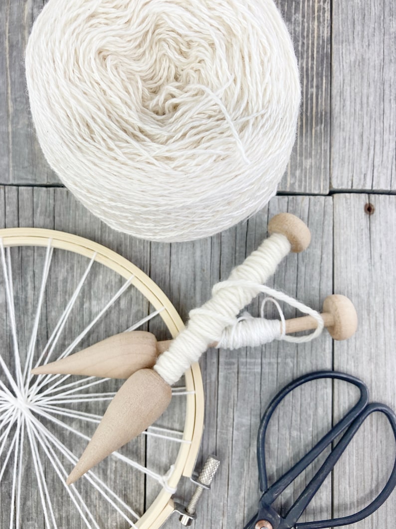 May include: A ball of white yarn, a wooden loom with white yarn wrapped around it, two wooden weaving tools, and a pair of black scissors on a wooden surface.