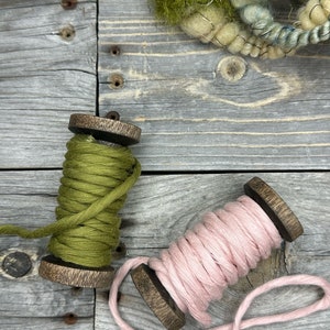 May include: Two spools of yarn, one green and one pink, are on a wooden surface. The yarn is wound around wooden spools.