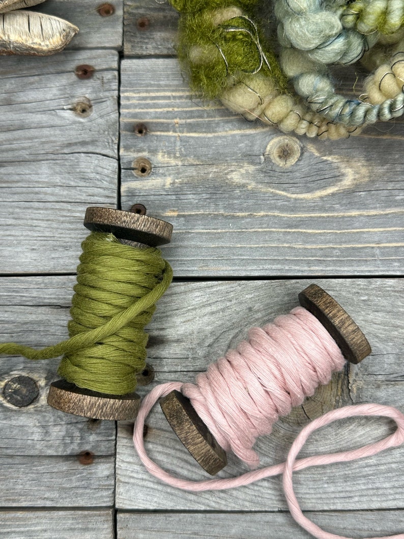 May include: Two spools of yarn, one green and one pink, are on a wooden surface. The yarn is wound around wooden spools.