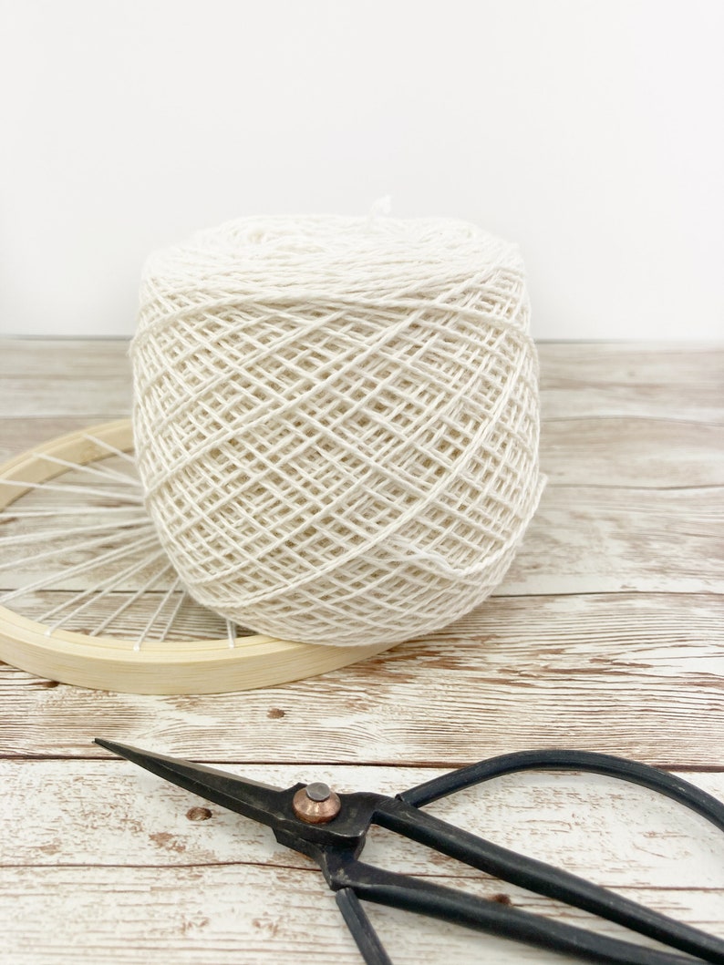 May include: A ball of white yarn sits on a wooden surface next to a wooden embroidery hoop and a pair of black metal scissors.