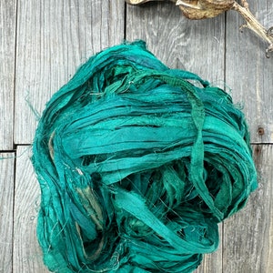 May include: A bundle of teal blue silk ribbon, also known as silk ribbon yarn, is shown on a wooden surface. The ribbon is made of thin strips of silk that have been twisted together.